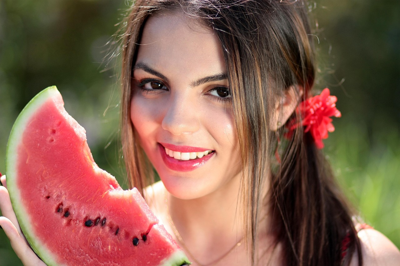 Woman holding and smiling while eating a fresh slice of watermelon outdoors on a sunny summer day, representing a healthy hydrating snack