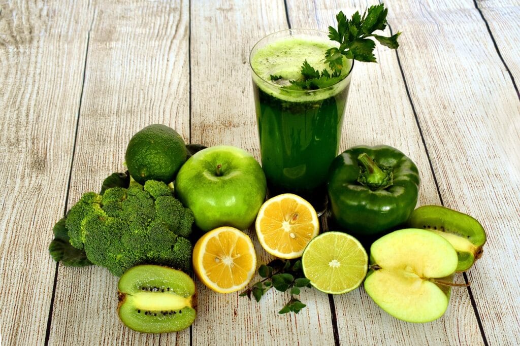Vibrant green juice smoothie surrounded by fresh fruits (apples, kiwi, lemon, lime) and vegetables (broccoli, bell pepper) on a light background, representing healthy natural drinks