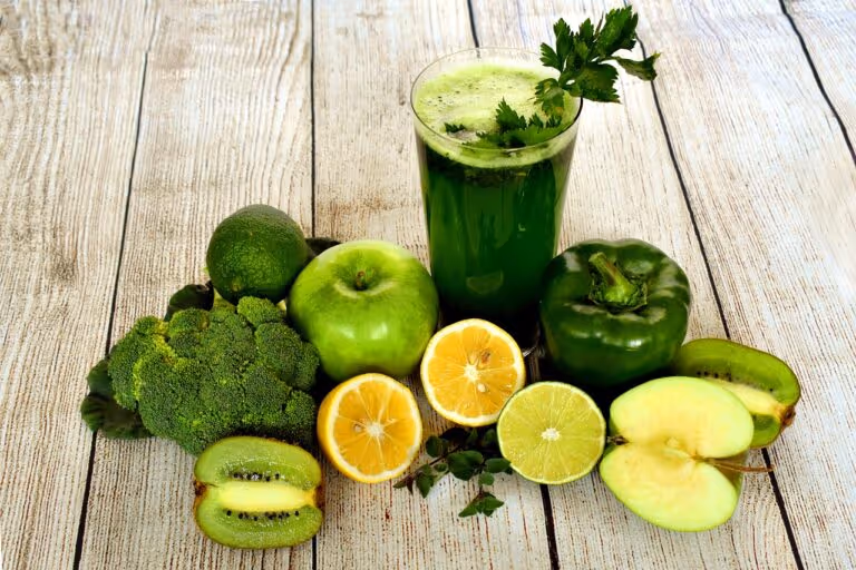 Vibrant green juice smoothie surrounded by fresh fruits (apples, kiwi, lemon, lime) and vegetables (broccoli, bell pepper) on a light background, representing healthy natural drinks