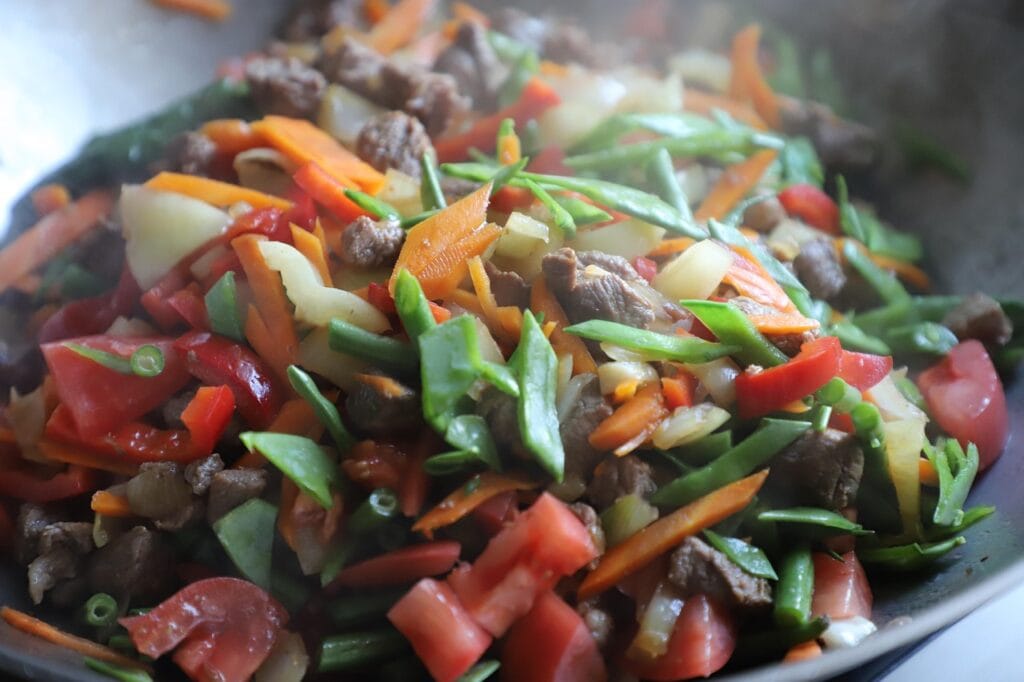 A close-up view of a colorful vegetable and meat stir-fry being cooked in a traditional wok, showing fresh broccoli, peppers, and other vegetables with minimal oil for healthy wok cooking.