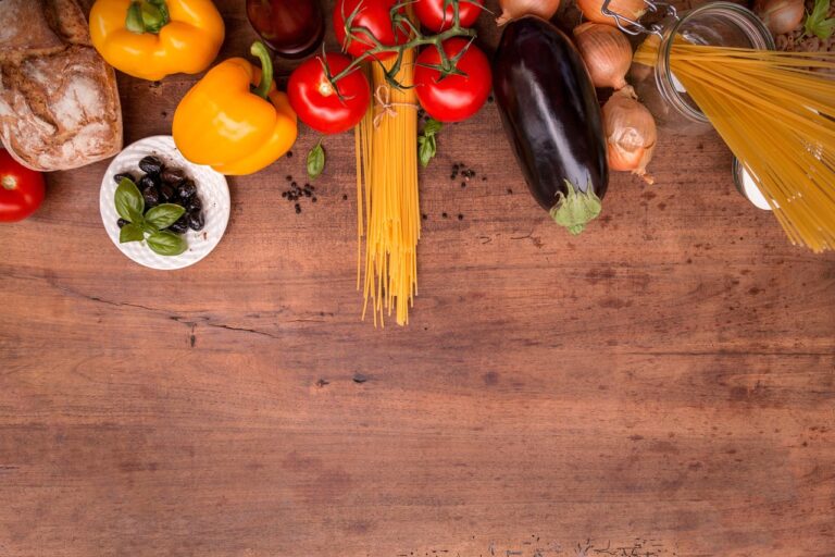 Fresh Mediterranean diet ingredients including vegetables, olives, pasta and herbs arranged on a wooden board, representing heart-healthy whole foods rich in healthy fats and nutrients