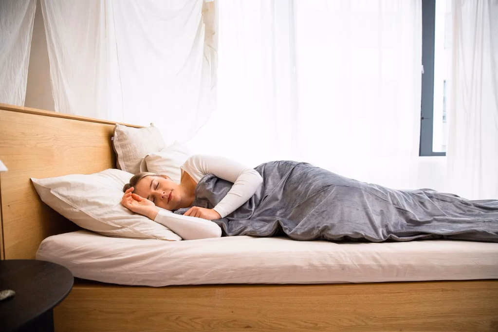 Woman sleeping peacefully on a non-toxic organic mattress in a bright, natural bedroom with wood tones, representing healthy sleep and wellness