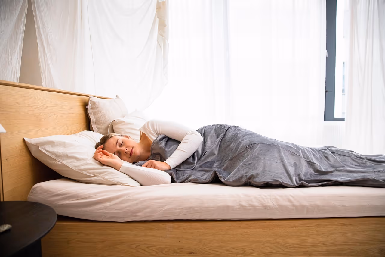 Woman sleeping peacefully on a non-toxic organic mattress in a bright, natural bedroom with wood tones, representing healthy sleep and wellness