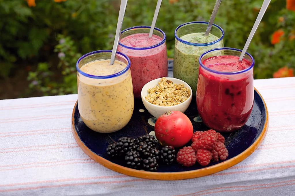 Colorful smoothies in glasses on a table, with fresh blueberries, raspberries, and granola nearby — perfect for a high fiber smoothie breakfast with banana, blueberry, and chia seeds.