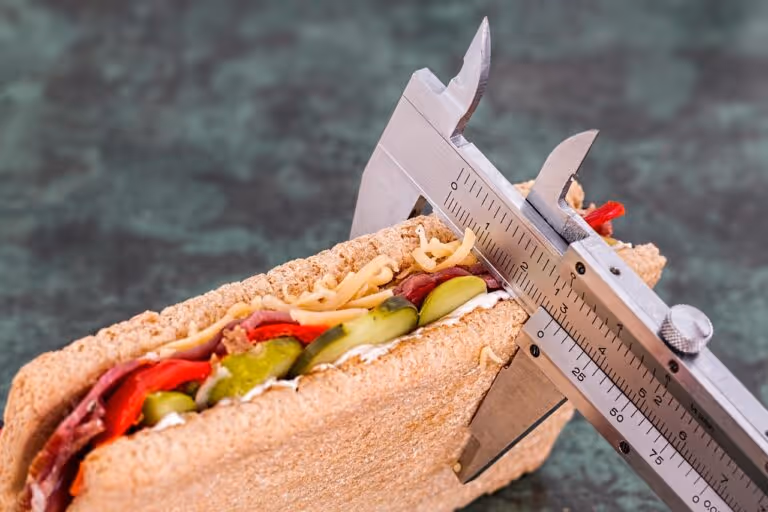 Close-up of a hand holding a vernier caliper measuring a sandwich, representing diet tracking, portion control, and weight loss management