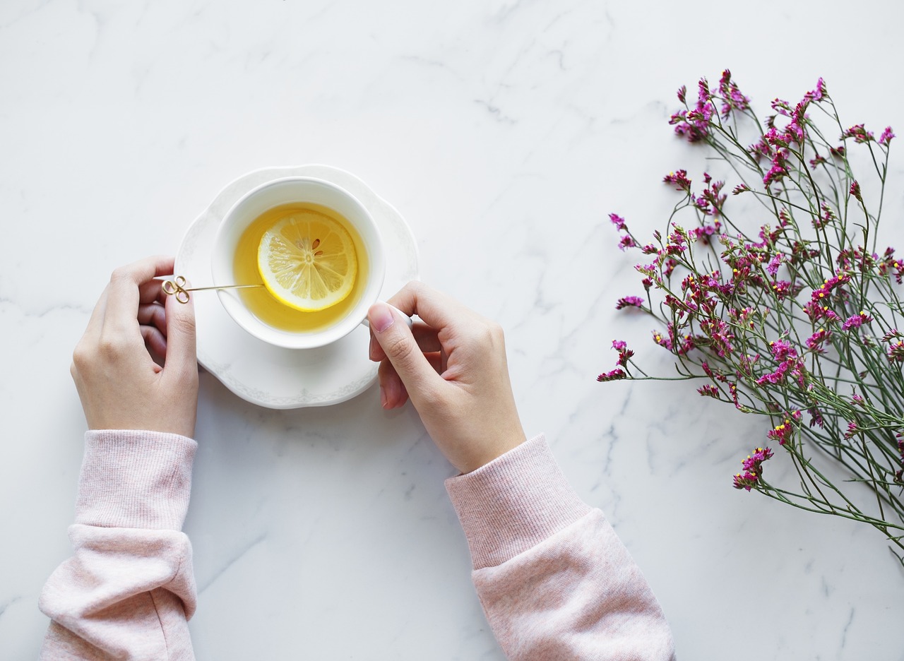 A cozy flatlay of hands holding a warm cup of tea with lemon slices and floral decorations, representing the mood-boosting ritual of pausing to enjoy a beverage
