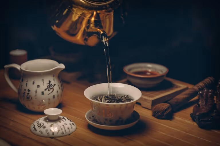 Close-up of a ceramic tea cup being filled with brewed green tea from a traditional teapot, with steam rising and scattered tea leaves on a wooden surface, representing natural metabolic health support