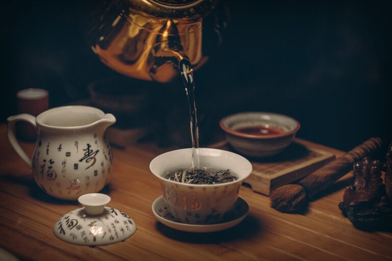 Close-up of a ceramic tea cup being filled with brewed green tea from a traditional teapot, with steam rising and scattered tea leaves on a wooden surface, representing natural metabolic health support