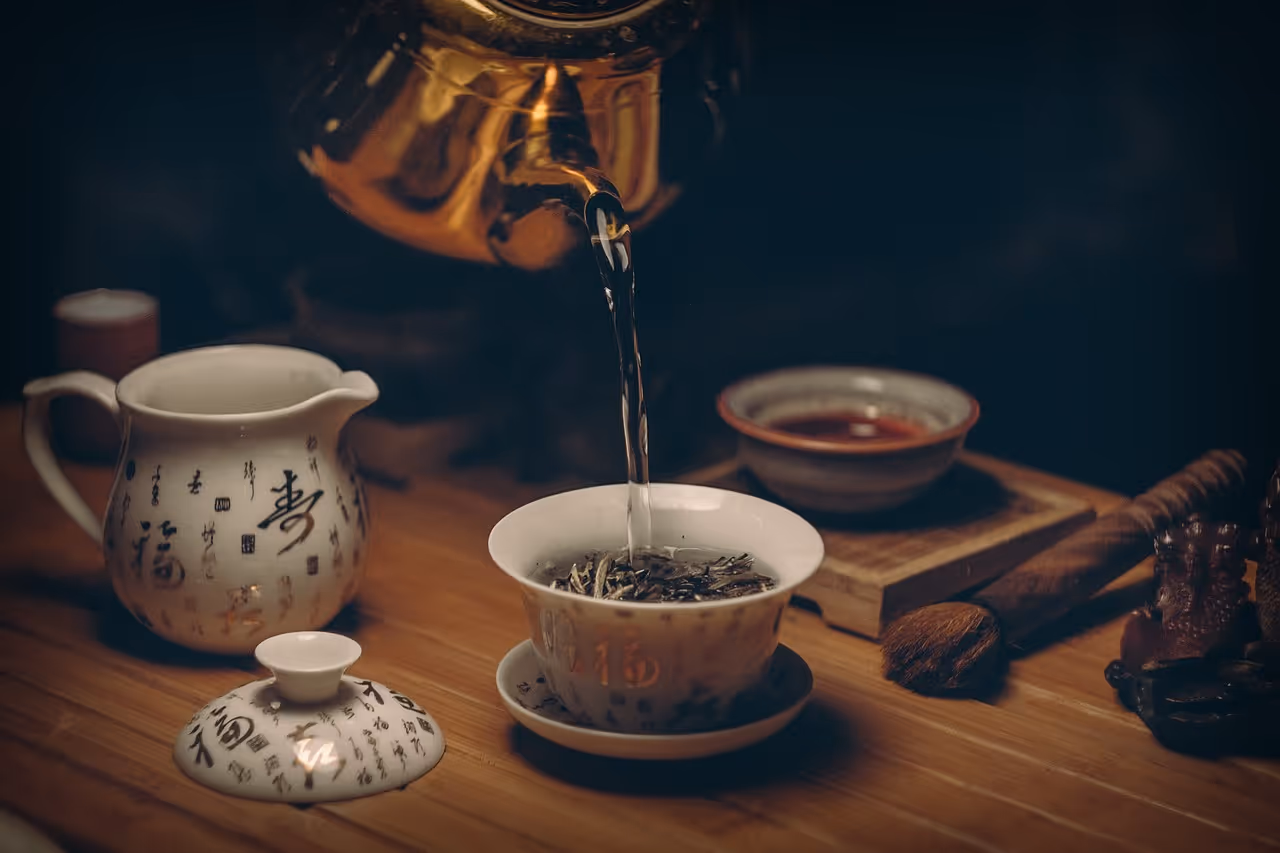 Close-up of a ceramic tea cup being filled with brewed green tea from a traditional teapot, with steam rising and scattered tea leaves on a wooden surface, representing natural metabolic health support
