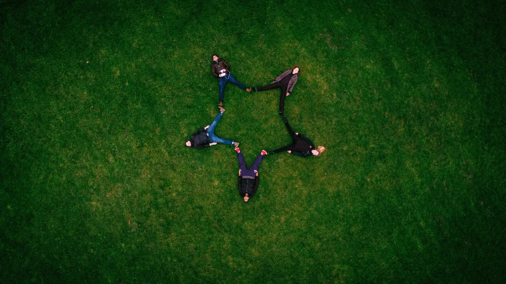 A group of adults lying on green grass in a field, forming a star shape formation with their bodies, symbolizing friendship, connection, and community outdoors under natural sunlight