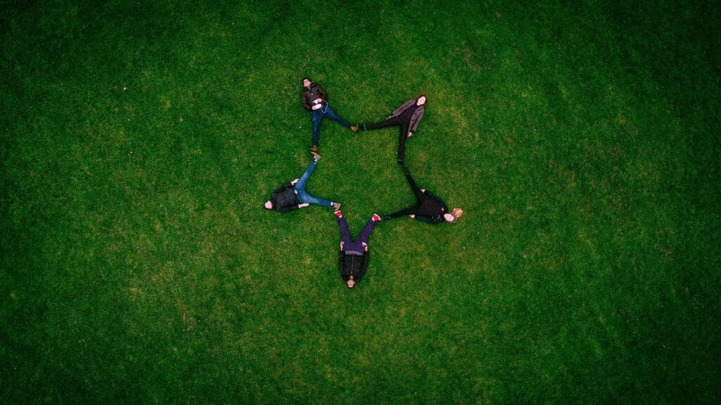 A group of adults lying on green grass in a field, forming a star shape formation with their bodies, symbolizing friendship, connection, and community outdoors under natural sunlight