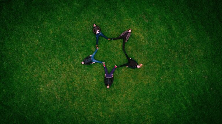 A group of adults lying on green grass in a field, forming a star shape formation with their bodies, symbolizing friendship, connection, and community outdoors under natural sunlight