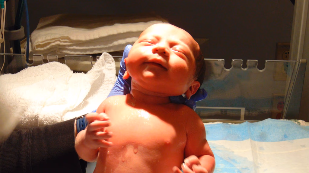 Newborn baby lying in a hospital bassinet, wrapped in a blanket, with a nurse or doctor nearby - representing newborn care and the vitamin K shot administered shortly after birth