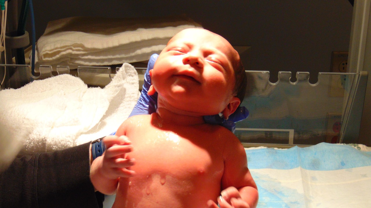 Newborn baby lying in a hospital bassinet, wrapped in a blanket, with a nurse or doctor nearby - representing newborn care and the vitamin K shot administered shortly after birth