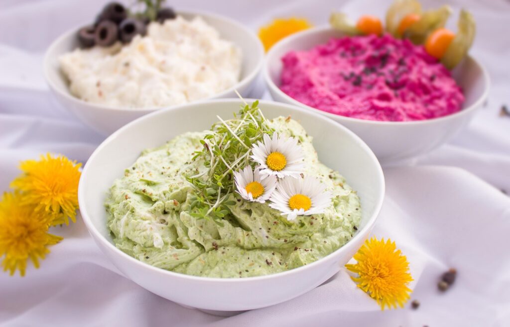 Three bowls of appetizer dips and spreads on a wooden table surrounded by flowers and fresh ingredients, perfect for quick summer appetizers and healthy entertaining.