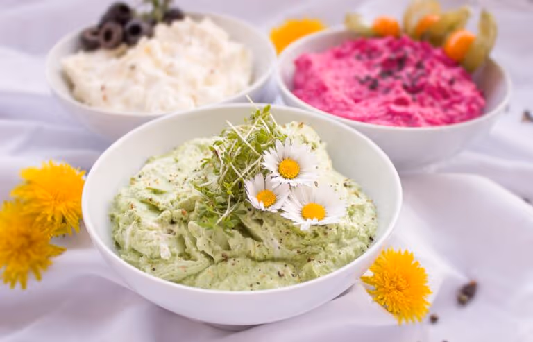 Three bowls of appetizer dips and spreads on a wooden table surrounded by flowers and fresh ingredients, perfect for quick summer appetizers and healthy entertaining.