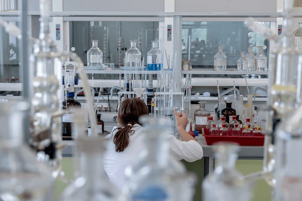 A female scientist wearing safety goggles and a lab coat working in a laboratory, representing cancer genetics research into the PDZRN4 gene that may explain why some tumors never become aggressive