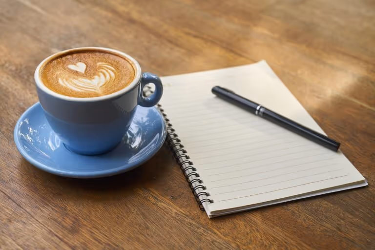 A cup of coffee with latte art on a table next to an open notebook and pen, representing the concept of pre-workout coffee as a natural performance booster for active women