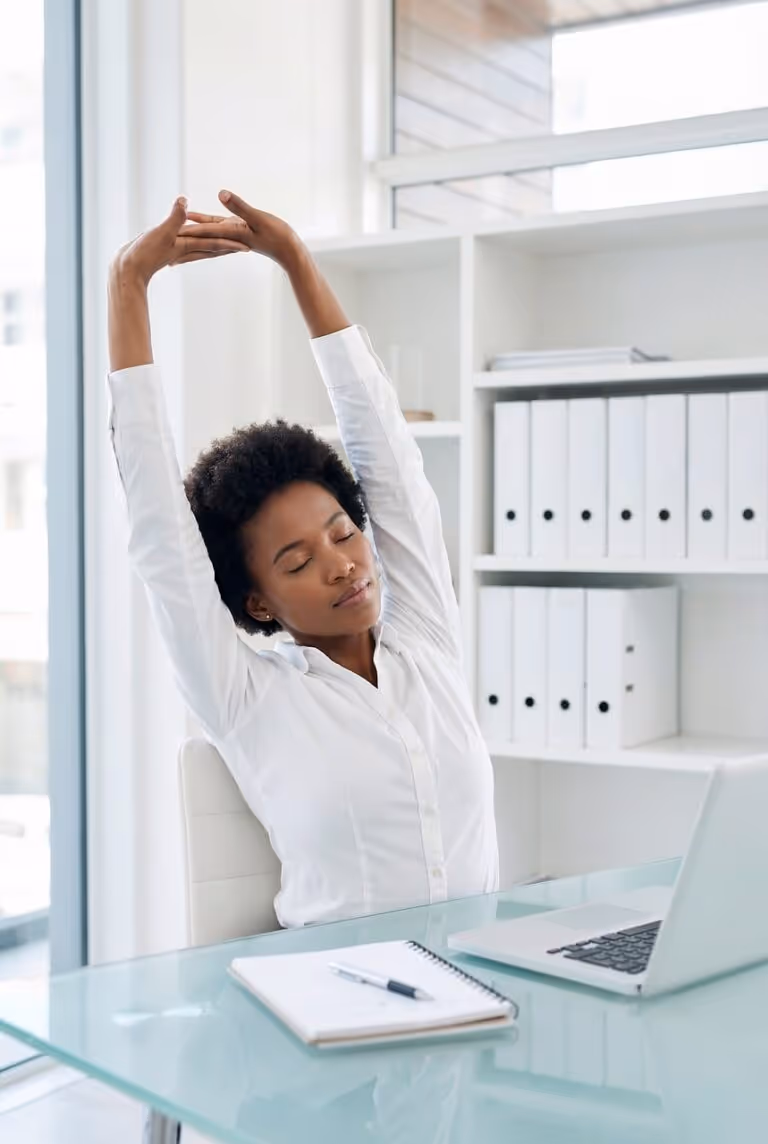 Woman in an office stretching her arms while sitting at a desk with a laptop, illustrating posture-improving movement breaks