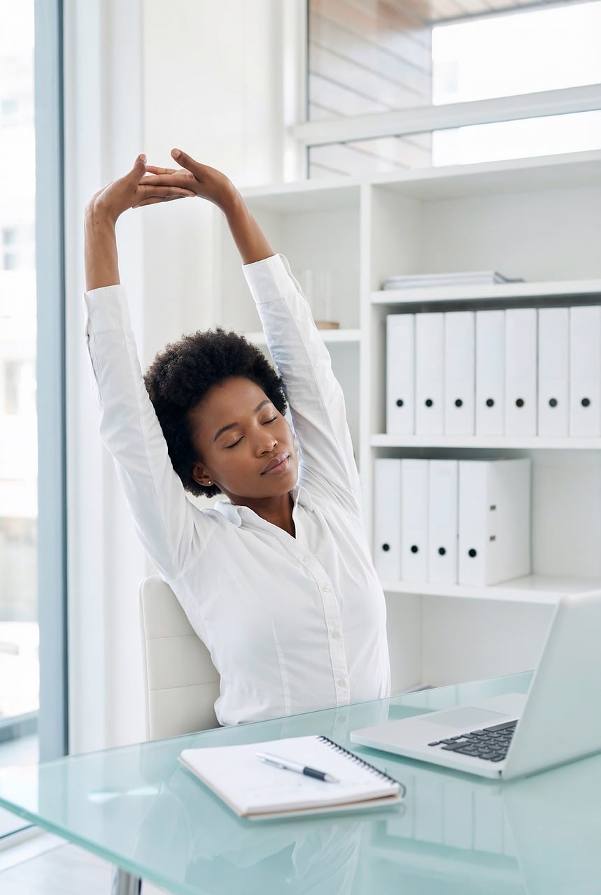 Woman in an office stretching her arms while sitting at a desk with a laptop, illustrating posture-improving movement breaks