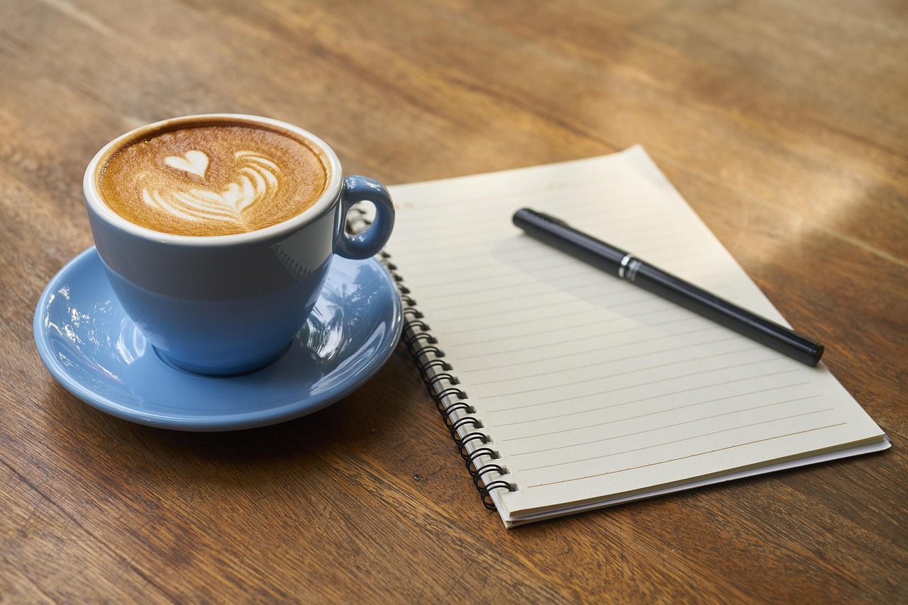 A cup of coffee with latte art resting on an open notebook, representing the morning ritual of enhancing coffee with healthy additives like cinnamon, cacao, and collagen