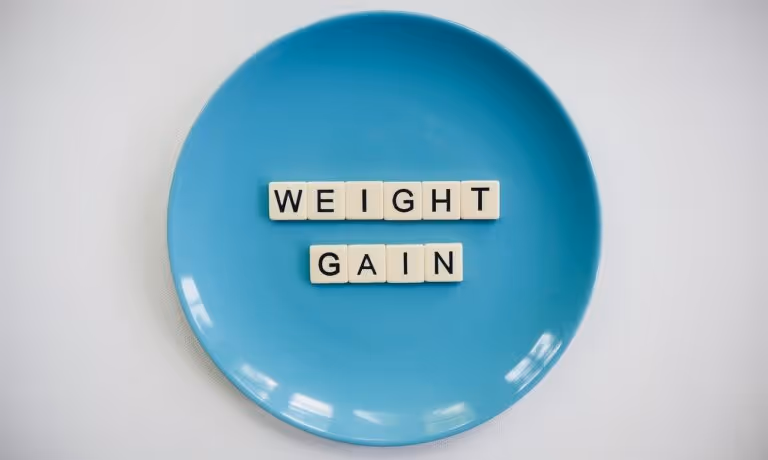 Scrabble tiles spelling out WEIGHT GAIN on a blue plate, representing the connection between sleep loss and weight gain from a 2026 study.