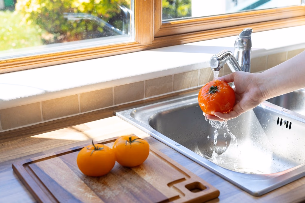 Hand washing a fresh tomato under running water in a kitchen sink, illustrating food safety and produce hygiene practices recommended during the cyclosporiasis outbreak