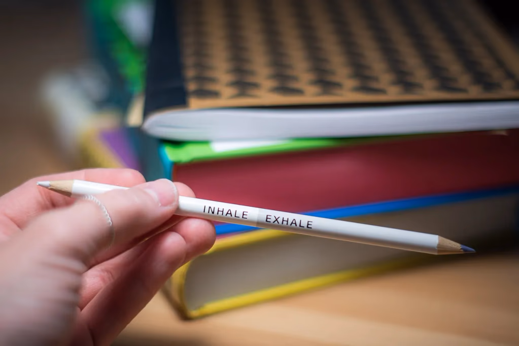 A pencil with 'INHALE EXHALE' written on it rests on a notebook on a wooden desk, with books in the background, illustrating the concept of longer exhale breathing for stress relief and nervous system calm.