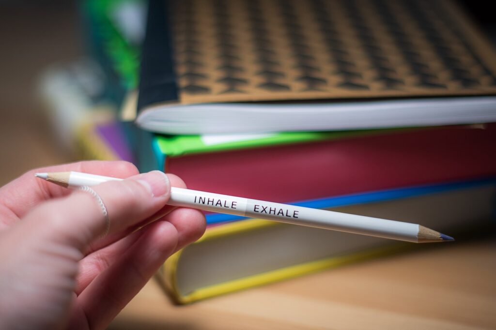 A pencil with 'INHALE EXHALE' written on it rests on a notebook on a wooden desk, with books in the background, illustrating the concept of longer exhale breathing for stress relief and nervous system calm.