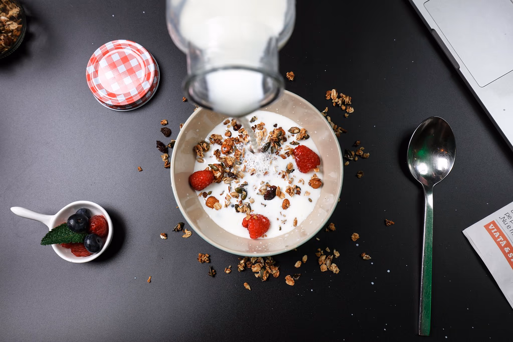 A bowl of Greek yogurt with fresh berries and granola, with milk or cream being poured over it, set on a wooden table in natural morning light