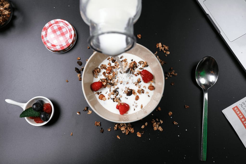 A bowl of Greek yogurt with fresh berries and granola, with milk or cream being poured over it, set on a wooden table in natural morning light