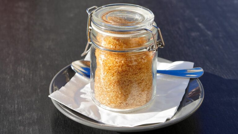 Glass jar filled with cane sugar cubes next to a coffee cup on a wooden table, representing hidden sugar in coffee shop drinks