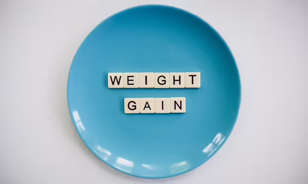Scrabble tiles spelling out WEIGHT GAIN on a blue plate, representing the connection between sleep loss and weight gain from a 2026 study.