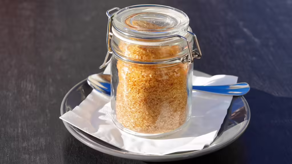 Glass jar filled with cane sugar cubes next to a coffee cup on a wooden table, representing hidden sugar in coffee shop drinks